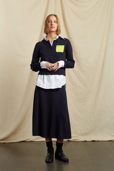 Woman wearing a navy shirt with a white collar and a yellow pocket square, standing against a beige curtain.