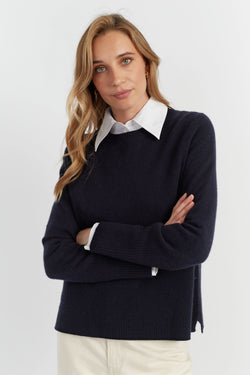 Woman wearing a navy sweater over a white shirt with arms crossed on a light gray background