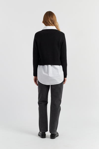 Person wearing a black sweater over a white shirt with gray pants on a white background