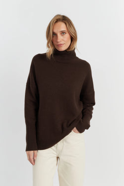 Woman wearing a brown turtleneck sweater and white pants on a light gray background