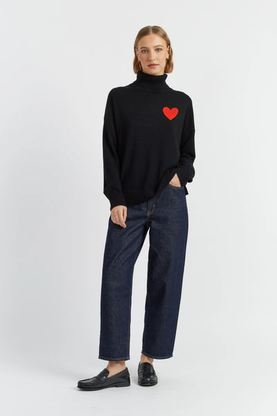 Person wearing a black turtleneck with a red heart design and blue jeans on a white background