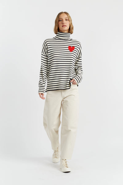 Woman wearing a black and white striped sweater with a red heart design, paired with white pants and shoes on a white background.