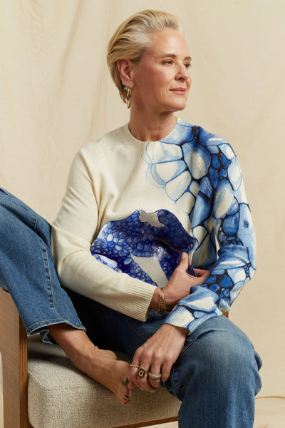 Woman wearing a cream sweater with blue floral pattern sitting on a beige chair.