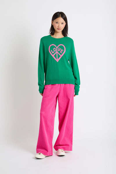 Person wearing a green sweater with a pink heart design and pink pants on a white background